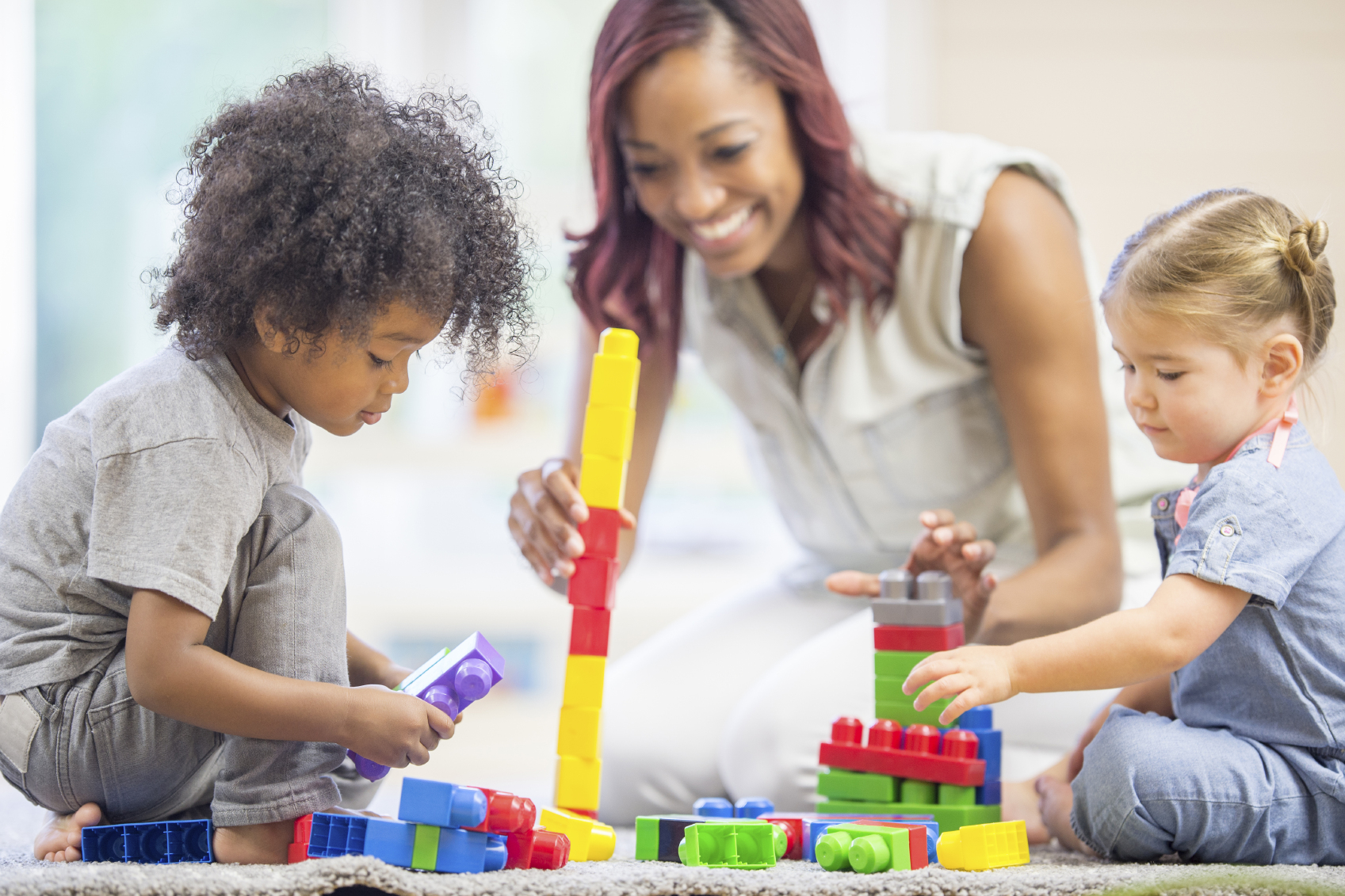 A multi-ethnic group of toddlers are playing with blocks on the floor with their teacher.